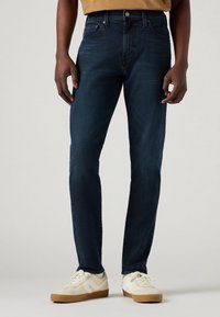 Mörkblå denimjeans med slim fit, som har en klassisk femfickdesign och en subtil textur. Kombineras med beige och vita sneakers.