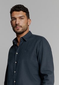Donkerblauwe knoopshirt met een spread collar, lange mouwen en contrasterende knoopdetails; gemaakt van een gestructureerde stof.
