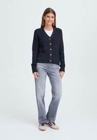 Cardigan en tricot bleu marine avec un col en V, doté de cinq grands boutons, associé à un jean gris clair et des chaussures décontractées, se tenant devant un fond clair.