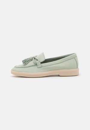 Mocassins en daim vert avec un nœud et des détails de glands sur le bout, dotés d'une semelle en caoutchouc texturée beige clair.