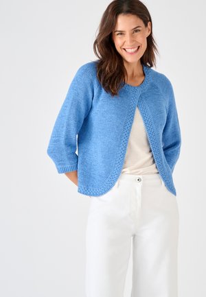 Femme souriante portant un cardigan en maille bleu à manches trois-quarts par-dessus un haut beige et un pantalon blanc sur un fond clair uni.