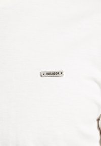 Hvid bomulds T-shirt med et lille sølvmetal mærke med "SMILODOX" indgraveret. Glat tekstur med et klassisk rundhalset design.