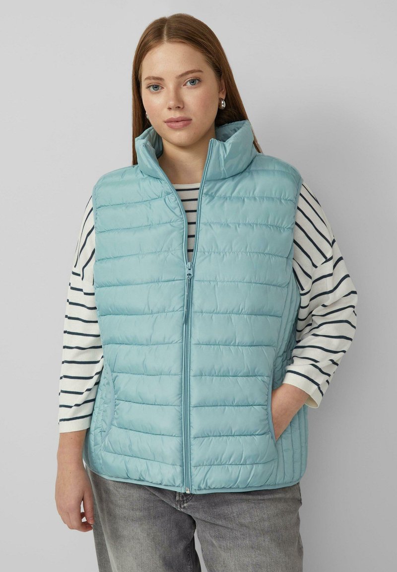 Lichtblauwe gewatteerde bodywarmer met opstaande horizontale naden, hoge kraag en zijzakken. Draagt over een gestreept shirt met lange mouwen.