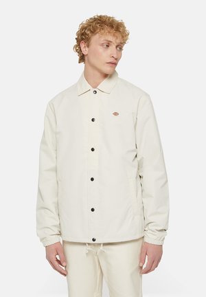 Chaqueta de botón en color beige claro, con cuello, puños elásticos y bolsillos laterales. Presenta un pequeño logo en el lado izquierdo del pecho. Textura suave.