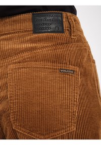 Brune corduroy bukser med lodrette ribber, et sort tekstureret logo-mærke på bagsiden og en synlig lomme med syninger.