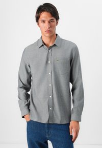 Camisa de botones gris con tejido texturizado, mangas largas y un logo verde en el bolsillo del pecho. Llevada con jeans azules.