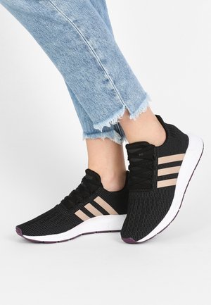 Personne portant un jean bleu clair effiloché et des baskets noires avec des bandes beiges et des semelles blanches, debout sur une surface blanche.