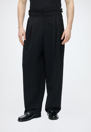 Mand iført løstsiddende sorte plisserede bukser med sidespænde og sorte læder loafers mod en hvid baggrund.