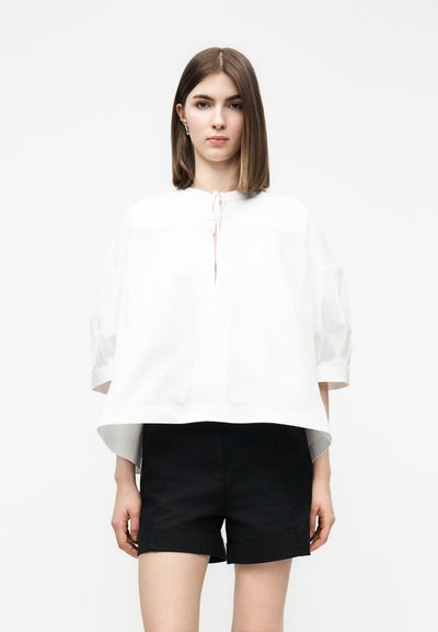 Jeune femme portant un chemisier blanc ample avec un petit nœud à l'avant et un short noir arrivant aux genoux, debout devant un fond uni.