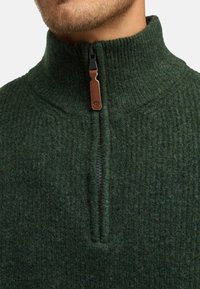 Dypgrønn fleece-pullover med høyt krage, med en delvis glidelås og en brun lær-glidelåsoppdraget. Teksturert overflate med ribbing.