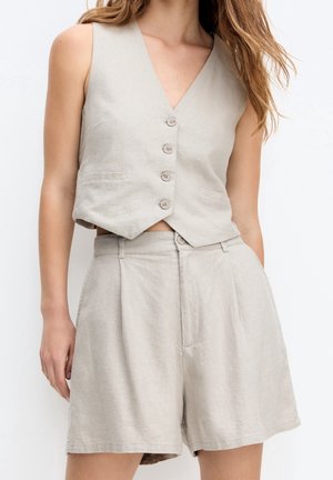 Vrouw draagt beige mouwloos gilet met knopen en bijpassende losse shorts met hoge taille tegen een witte achtergrond.