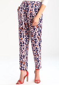 Pantalons à motifs géométriques en rose, bleu marine et orange. Tissu léger, coupe ajustée avec poches latérales et finitions aux chevilles. Associés à des talons hauts rouges.