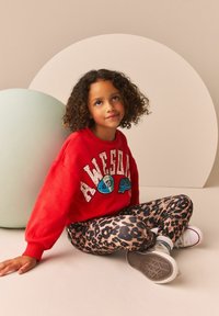 Un sweat-shirt rouge avec un graphisme "SUPER", associé à des leggings à imprimé léopard. Un enfant est assis contre une forme ronde vert clair. Des baskets blanches.