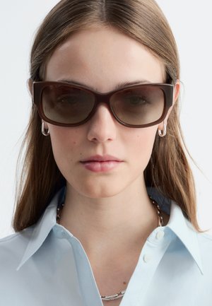 Kobieta o prostych brązowych włosach nosi duże ciemnobrązowe okulary przeciwsłoneczne, srebrne kolczyki-koła, srebrny naszyjnik na łańcuszku oraz jasnoniebieską koszulę z kołnierzykiem.