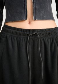 Pantalons de jogging en polaire noire avec une taille élastique, dotés d'un détail à cordon de serrage. Le haut est un sweat à zip en velours côtelé noir, court.
