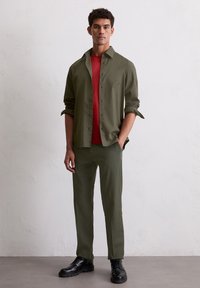 Olijfgroene knoopshirt over een rode t-shirt, gecombineerd met bijpassende olijfgroene broek en zwarte schoenen. Glad materiaal, casual pasvorm.