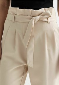 Beige hoge taille broek met paperbag taille, frontplooien, riemlussen en een gestrikt stoffen riem, gedragen met een zwarte top.