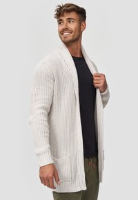 Cardigan a maglia beige chiaro con apertura frontale, polsini a coste e tasche laterali. Tessuto intrecciato, vestibilità rilassata, indossato sopra una camicia nera.