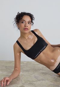 Čierny bralet s širokými ramienkami a logom na páse; hladký materiál; model leží na vzorovanej podlahe s viditeľnými variáciami pleti.