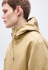 Veste imperméable beige avec un col montant, une capuche ajustable et un cordon de serrage. Présente un discret logo « cK » embossé sur la manche gauche.