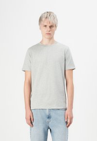Tânăr cu părul blond scurt, purtând un tricou simplu gri deschis și blugi albaștri deschiși, stând pe un fundal alb.