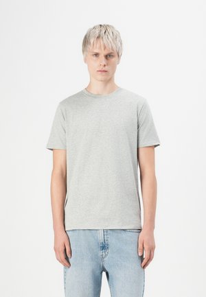 Junge Person mit kurzen blonden Haaren, die ein schlichtes, hellgraues T-Shirt und hellblaue Jeans trägt, steht vor einem weißen Hintergrund.