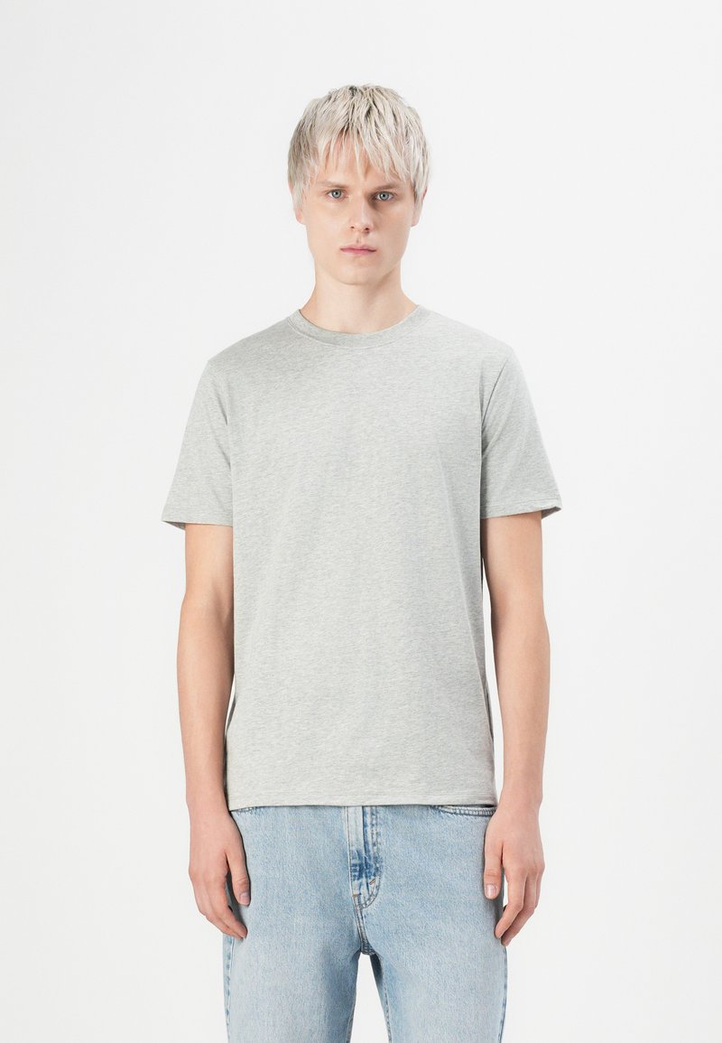 Tânăr cu părul blond scurt, purtând un tricou simplu gri deschis și blugi albaștri deschiși, stând pe un fundal alb.
