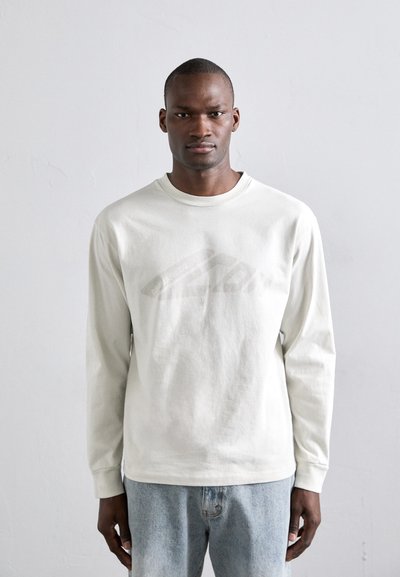 Camiseta de algodón gris claro de manga larga con un sutil logo tonal grande en la parte delantera, combinada con jeans de mezclilla azules.