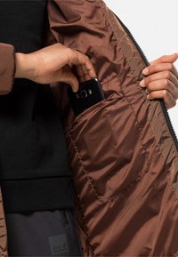 Bruine geïsoleerde jas met een glanzende binnenkant, met een zijzak waarin een zwarte smartphone zit. Draagt over een zwarte trui.