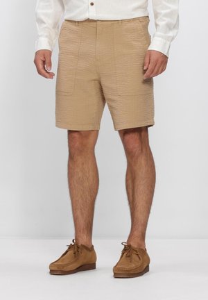 Hombre con pantalones cortos beige texturizados, zapatos marrones de ante con cordones y camisa blanca de manga larga, de pie sobre un fondo liso.
