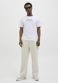 Hvid t-shirt med korte ærmer og sort tekst, løse cremefarvede bukser og grå sneakers med hvide detaljer, suppleret med mørke solbriller.