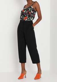 Kukkaroitu musta kamisol, jossa on säädettävät hihnat, yhdistettynä korkeavyötäröisiin mustiin leveälahkeisiin housuihin ja oransseihin muloihin, joissa on kultaiset yksityiskohdat.