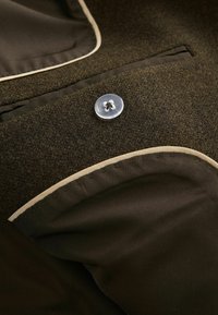 Hnědá vlněná látka s béžovými akcenty, obsahující průhledný knoflík a kapsu, vykazující hladkou texturu a přizpůsobený design.