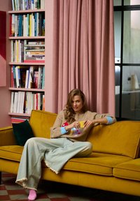 Żółta sofa z materiału z postrzępionymi krawędziami, różowe zasłony i drewniana półka w tle. Osoba trzyma żółty kubek, ubrana w beżowy sweter z kolorowymi wzorami.