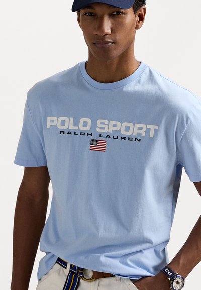 Világoskék pamut póló, fehér szöveggel "POLO SPORT" és "RALPH LAUREN", kicsi amerikai zászló grafikával. Rövid ujjú és kerek nyakú.