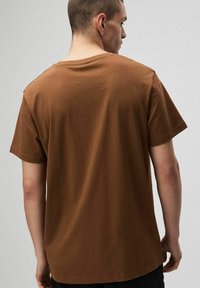 Mężczyzna odwrócony tyłem, ubrany w prostą brązową koszulkę z krótkim rękawem i okrągłym dekoltem oraz czarne spodnie, na neutralnym tle.