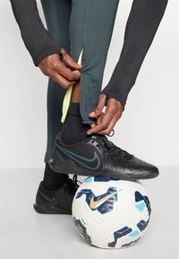 Crne Nike nogometne tenisice na bijeloj i plavoj uzorcima lopti. Osoba podešava zelene atletske hlače s detaljima žutog patentnog zatvarača.