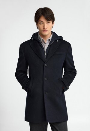 Uomo che indossa un cappotto nero con bottoni, una camicia grigia con colletto e una felpa nera con zip sotto, mani nelle tasche del cappotto.
