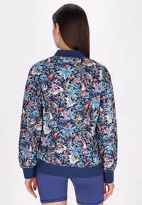 Giacca bomber floreale in blu navy con vivaci motivi di fiori tropicali e uccelli. Polsini e orlo a coste. Tessuto morbido e testurizzato.