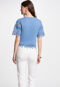 Blå kortærmet top med blondedetaljer ved ærmerne og kanten, parret med hvide lige ben jeans. Stoffet ser glat ud.