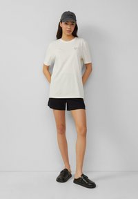 T-shirt en coton blanc à manches courtes, shorts noirs et chaussures noires à enfiler. La casquette est grise avec du texte noir. Le modèle se tient devant un fond gris.