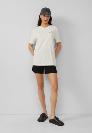 Biała bawełniana koszulka z krótkim rękawem, czarne szorty i czarne buty typu slip-on. Czapka jest szara z czarnym napisem. Model stoi na szarym tle.
