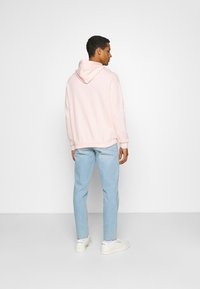 Lys rosa hettegenser med snor i hetten, avslappet passform, ribbede mansjetter, sammen med lyseblå jeans og hvite sneakers. Ensfarget bakgrunn.