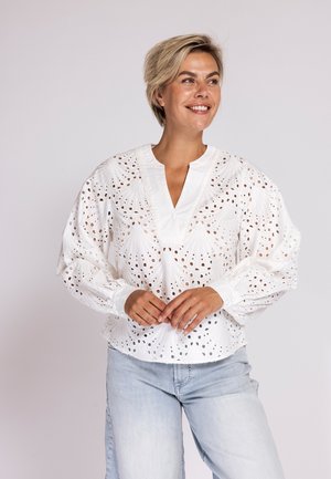 Lächelnde Frau mit kurzen blonden Haaren, die eine weiße Bluse mit Lochmuster und langen Ärmeln sowie hellblaue Jeans vor einem schlichten Hintergrund trägt.