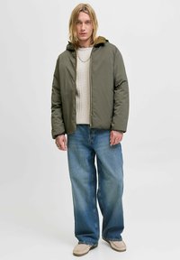 Olivgrüne gepolsterte Jacke mit Kapuze, Reißverschluss und darunter einem ribbelstrick Pullover. Kombiniert mit lockeren blauen Jeans und beigen Slippern.