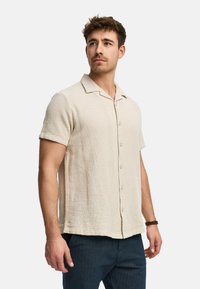Kortærmet skjorte med knapper i beige tekstureret stof med krave, der har en lige nederkant og knapper foran.