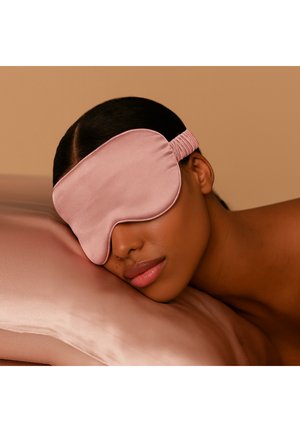 Różowa satynowa maska do spania o zakrzywionym kształcie i regulowanym elastycznym pasku, położona na gładkiej, jasnej poduszce. Miękka tekstura.