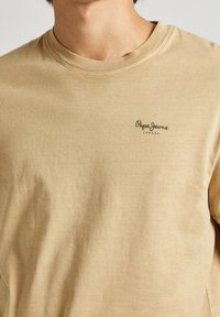 Kaki t-shirt i bomull med rund hals. Har en grön "Pepe Jeans London"-logotyp på bröstet. Slät textur, avslappnad design.