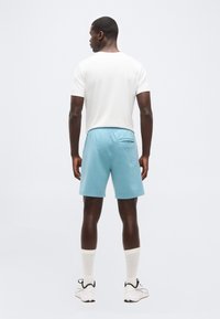 Άνδρας με την πλάτη γυρισμένη φοράει λευκό πουκάμισο με κοντά μανίκια, ανοιχτό μπλε σορτς με φερμουάρ στην πίσω τσέπη, λευκές κάλτσες και λευκά αθλητικά παπούτσια σε απλό φόντο.