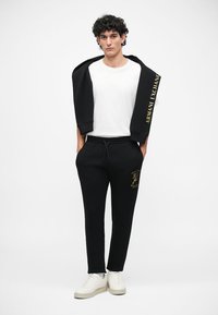 Mustarakk musta kapuutsiga särk, millel on 'Armani Exchange' logo, valge T-särk, mustad treeningpüksid kuldsete detailidega ja valged tossud, seismas ühtlase tausta ees.